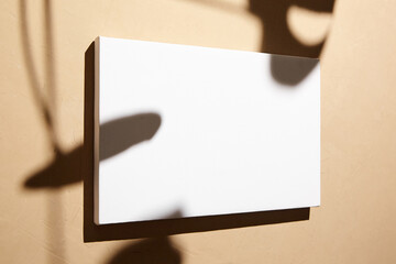 White canvas, blank picture mockup hanging on beige wall with dark shadows of leaves. Poster mockup, empty canvas with shadows of plant, side view