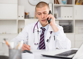 Male doctor working in the clinic consults a patient on the phone, sitting at a desk in the office