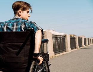 A young disabled man rides in a wheelchair across a bridge, the young male dressed in a plaid shirt...