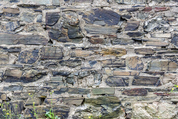 A closeup of an old building wall made from stone and mortar. A variety of rough stone makes up the wall. Bits of grass at the bottom.