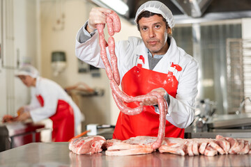 Portrait of positive professional butcher in white uniform and red apron holding chain of raw sausages while standing in processing workshop near fresh meat cutting table - Powered by Adobe