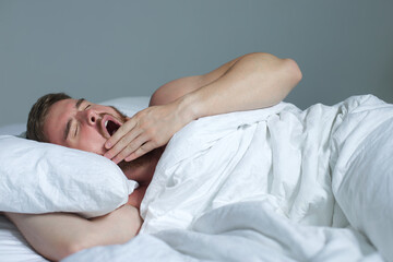 Young happy handsome sleepy man in the bed in bedroom at home in the morning lying under white blanket, enjoy resting, sleep well and yawn with opened mouth