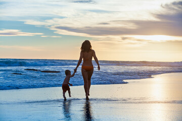 Fototapeta na wymiar Happy family - mother, baby son hold hands and run with fun along edge of sunset sea on sand beach. Active parents and people outdoor activity on tropical summer vacations with children