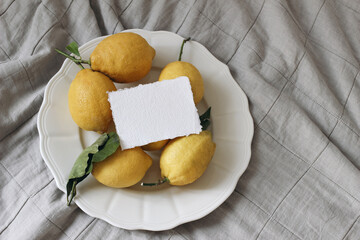Fresh yellow lemons, leaves on white ceramic plate. Business card mockup. Greige muslin tablecloth, fabric. Closeup of Mediterranean juicy fruit on table. Healthy eating. Flat lay, top view.