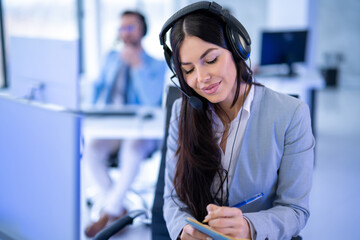 Portrait of attractive customer support service worker woman taking notes while listening customer over headset. Female operator at office. - obrazy, fototapety, plakaty