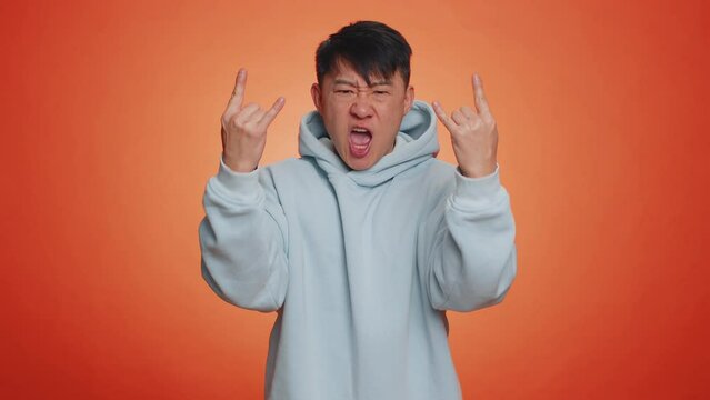 Overjoyed delighted asian handsome man showing rock n roll gesture by hands, cool sign, shouting yeah with crazy expression, dancing, emotionally rejoicing in success. Guy on orange studio background