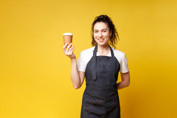girl barista in a denim apron holds cup of coffee and smiles on yellow background, portrait of...
