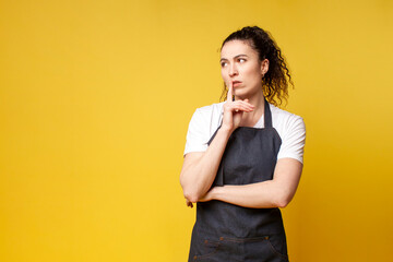 thoughtful young barista girl in uniform stands on a yellow background and thinks, a woman waiter...