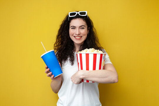 young girl brunette viewer in 3d glasses and white t-shirt holds large bucket of popcorn and a drink and smiles