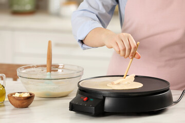Woman cooking delicious crepe on electric pancake maker at white marble table in kitchen, closeup