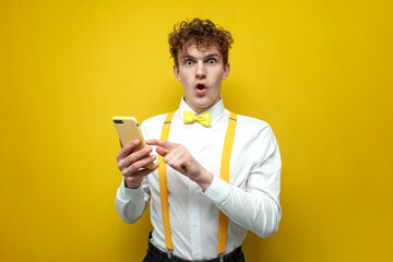 young curly guy in shirt with bow tie and suspenders holds smartphone and is surprised on yellow background