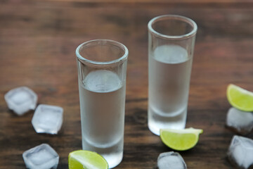 Mexican tequila shots with lime slices and ice cubes on wooden table, closeup. Drink made from agave
