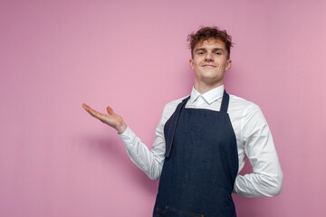 young waiter in a white shirt and an apron shows his hands on a copy space on a pink background
