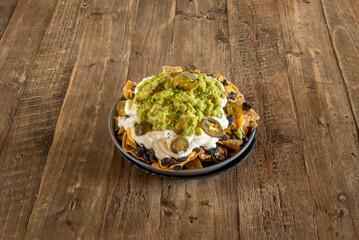 Round black clay bowl with nachos with melted cheese, pico de gallo and guacamole on unglazed wood table