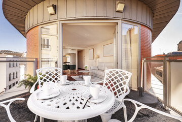Beautiful image of an urban terrace with a circular white outdoor table with matching chairs with breakfast cups and access to a living room with a white sofa