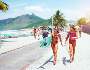 two young caucasian surfer women in swimsuits walking on the street with surfboards and talking to...