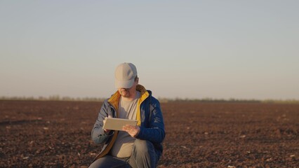 Man agronomist squats on black ground plot of plowed and cultivated field in countryside on spring day. Farmer enters data for report on soil condition into tablet. Agricultural business concept