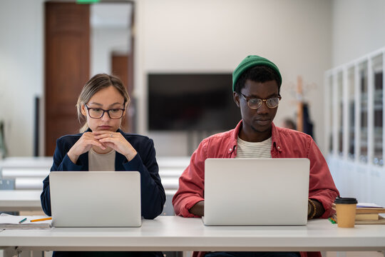 African American guy and Caucasian girl students in eyeglasses focused silently sits at desk looking laptops screen in university classroom. Multiethnic interns completing training task at workplace.