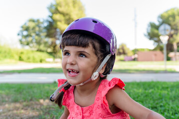 Fototapeta na wymiar Little girl looking at camera while wearing protective helmet outdoors in a park.