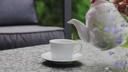 Close up of hand pouring black tea or coffee from ceramic teapot to a cup in a table. Summer outdoor background. Waiter service customer in caffe or restaurant terrace. High quality FullHD footage