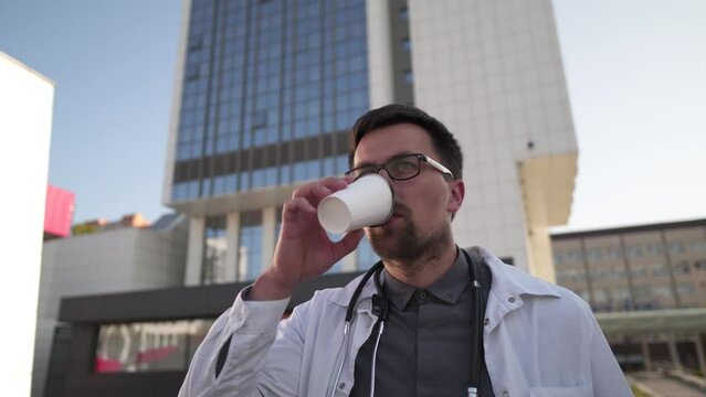 Medic in white medical coat goes and drinks take-out coffee outside hospital. Caucasian male young doctor during coffee break and walk in courtyard of clinic after hard shift. Medical profession.