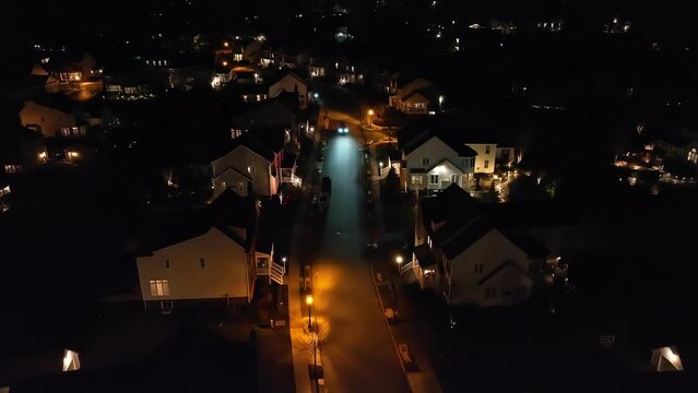 Aerial descending shot of car driving through dark American neighborhood. Upscale housing development in USA at night. Establishing shot of vehicle traveling with headlights at dark.