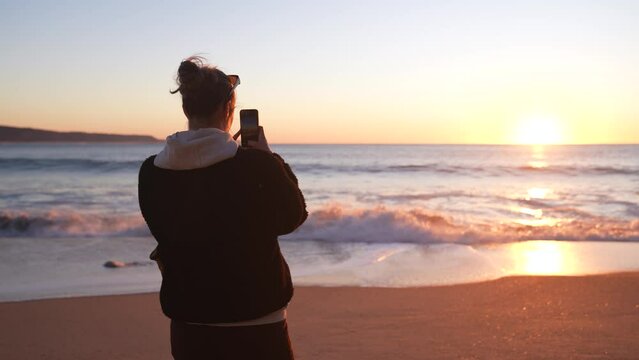 Young woman taking pictures with her smart phone on the sunset by the beach and wave break