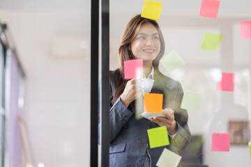 Asian businesswoman creating project plan on office wall with sticky paper notes. stylish confident manager working on business, financial and marketing planning projects.

