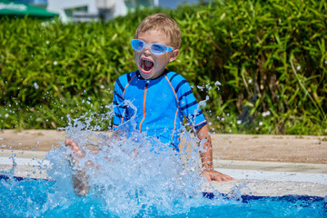 Little blonde boy baby splashes in the pool with his mouth wide open. Happy boy playing in the...
