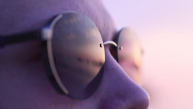 Young man in sunglasses looks at the sunset standing on a sandy beach by the sea.