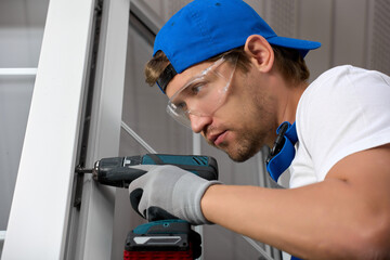 Focused male installer in blue cap and goggles holds screwdriver in his hand fixes mount on sash plastic window. Experienced master for installation plastic windows and doors with screwdriver in hand