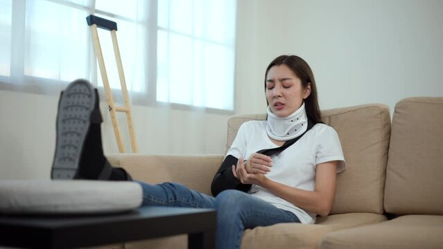 Woman suffered pain from accident fracture broken bone injury with leg splints in cast, neck splints collar, arm splints, sling support arm in living room. Social security and health insurance.