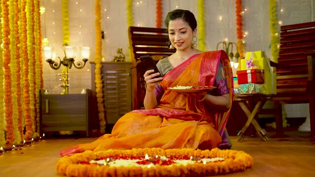 Happy young girl using mobile phone to check for new decorative designs by holding flowers plate during festival rangoli decoration at home - concept of technology, festive planning and social media