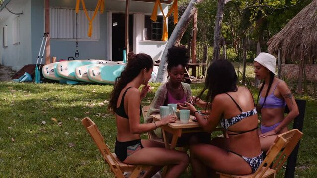 Four happy girls from different ethnic backgrounds share a meal around a table in a lush garden. Stand up paddle boards are visible in the back. Central America, backpacker vibes, travel images.