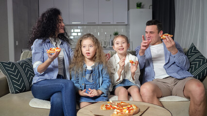 Family watches movies and enjoys eating yummy pizza together. Parents and daughters with amused and excited expression sit on sofa and watch comedy, closeup