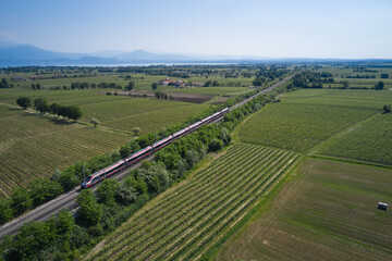 Fototapeta na wymiar Aerial view at high altitude gray train movement on the background of lake garda drone view. Gray high speed train moving in Blur in Italy vineyard fields.