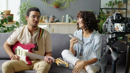 Gen Z couple sitting on sofa, talking and playing guitar and shaker while filming music blog with digital camera on tripod in living room at home