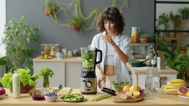Young woman putting fresh salad leaves into blender while cooking green smoothie in kitchen at home