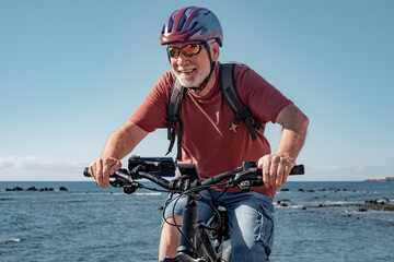 Cheerful caucasian senior man wearing helmet riding on the beach with electric bicycle. Authentic free and relaxed elderly lifestyle concept. Horizon over sea