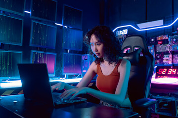 Female Asian hacker in hideout place with dark environment and multiple displays and cables, trying to break into data servers and infect the system with a virus. 