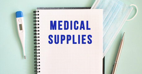 Notepad, medical mask, thermometer and pen on a blue background. MEDICAL SUPPLIES text in a notebook.