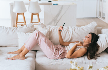 Obraz na płótnie Canvas Laughing Asian young brunette woman in pastel clothes laying on cozy couch at living room holds phone makes video call broad smiles. Excited girl relaxing at home on vacation. Domestic leisure.