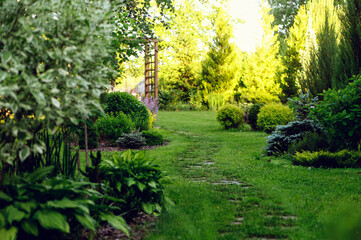 Fototapeta na wymiar Beautiful summer garden view with curvy stone pathway and wooden archway. Natural woodland cottage garden with hostas, conifers and shrubs
