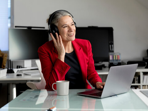 Happy Senior Businesswoman Wearing Wireless Headphones Sitting With Laptop At Desk