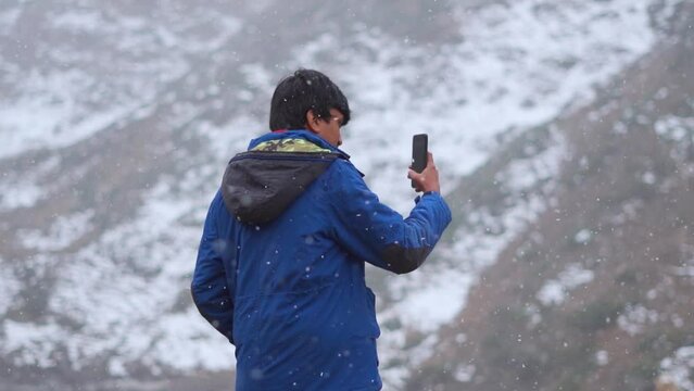 Slow motion shot of an Indian man shooting video of the snowfall with his mobile phone during the winter season at Sissu in Himachal Pradesh, India. Man taking video of snowfall for his social media.