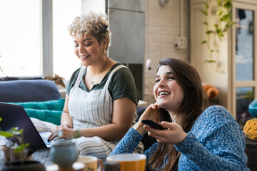 Happy young woman watching TV while friend using laptop at home