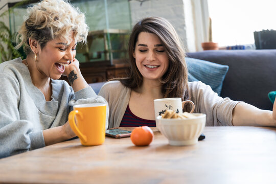 Happy female roommates using smart phone while having coffee at table