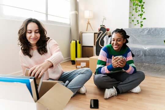 Smiling young woman unpacking box with friend holding coffee cup at home