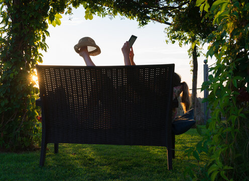 Backlighting Of A Blonde Woman Lying On A Wooden Bench Of A Garden With Her Head On A Cushion, Looking Something On A Tablet And Holding A Straw Hat With Her Foot Up At The Sunset.