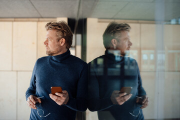 Thoughtful man with smart phone leaning on glass wall